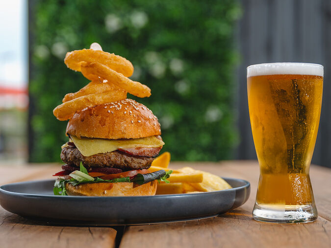 Juicy gourmet burger stacked with onion rings, crispy chips, and a cold beer — showcasing hearty, classic Caboolture food perfect for a laid-back local meal.